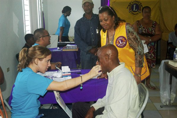 Photo by Valerie Dixon
Immediate Past President of the Lions Club of Mandeville Fay Heaven (right) observes a volunteer from the Miami Dade College Medical Group testing the eye of a patient during a recent wellness clinic in Manchester.
